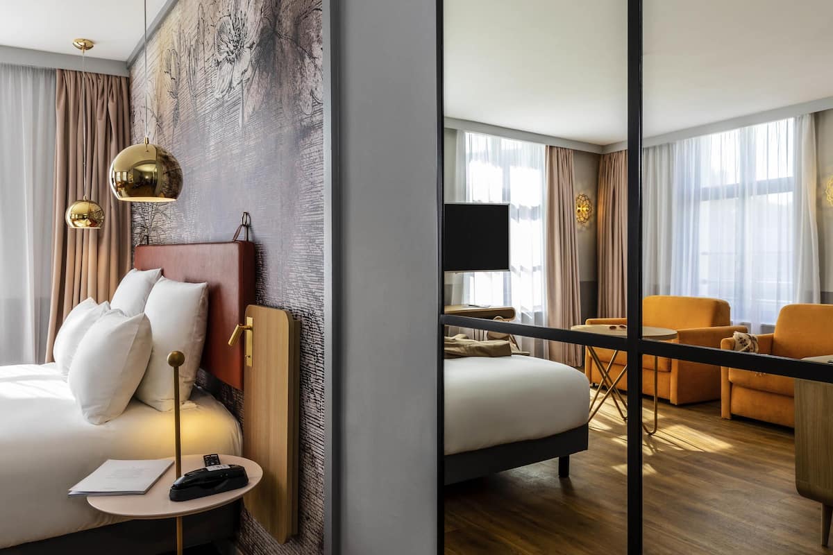 A hotel room with a bed, bedside table, lamp, and a large mirror reflecting the room.