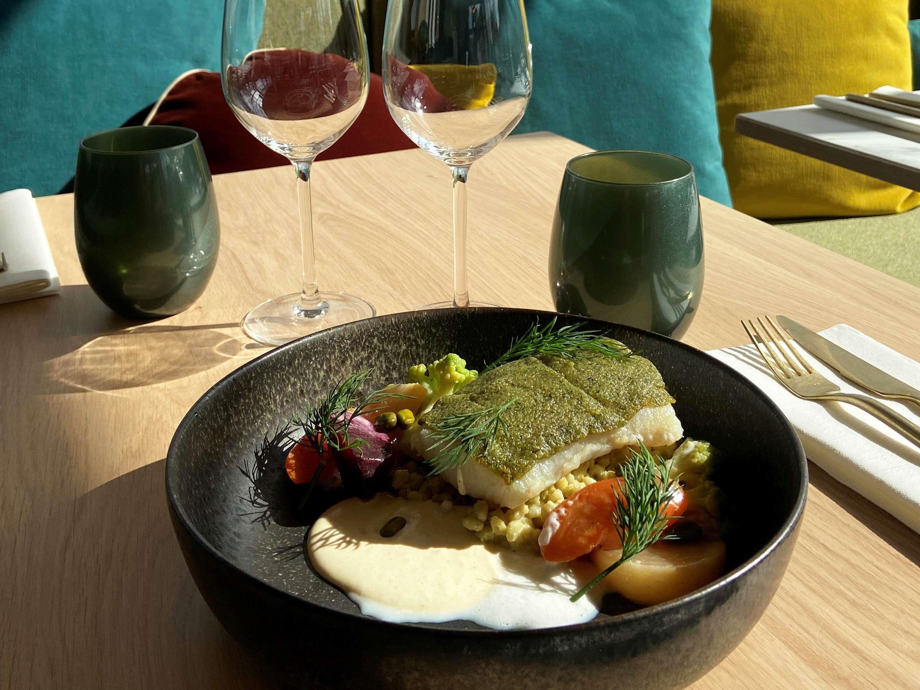 A dining table with a black bowl containing a fish dish, two wine glasses, and a fork and knife on a napkin.