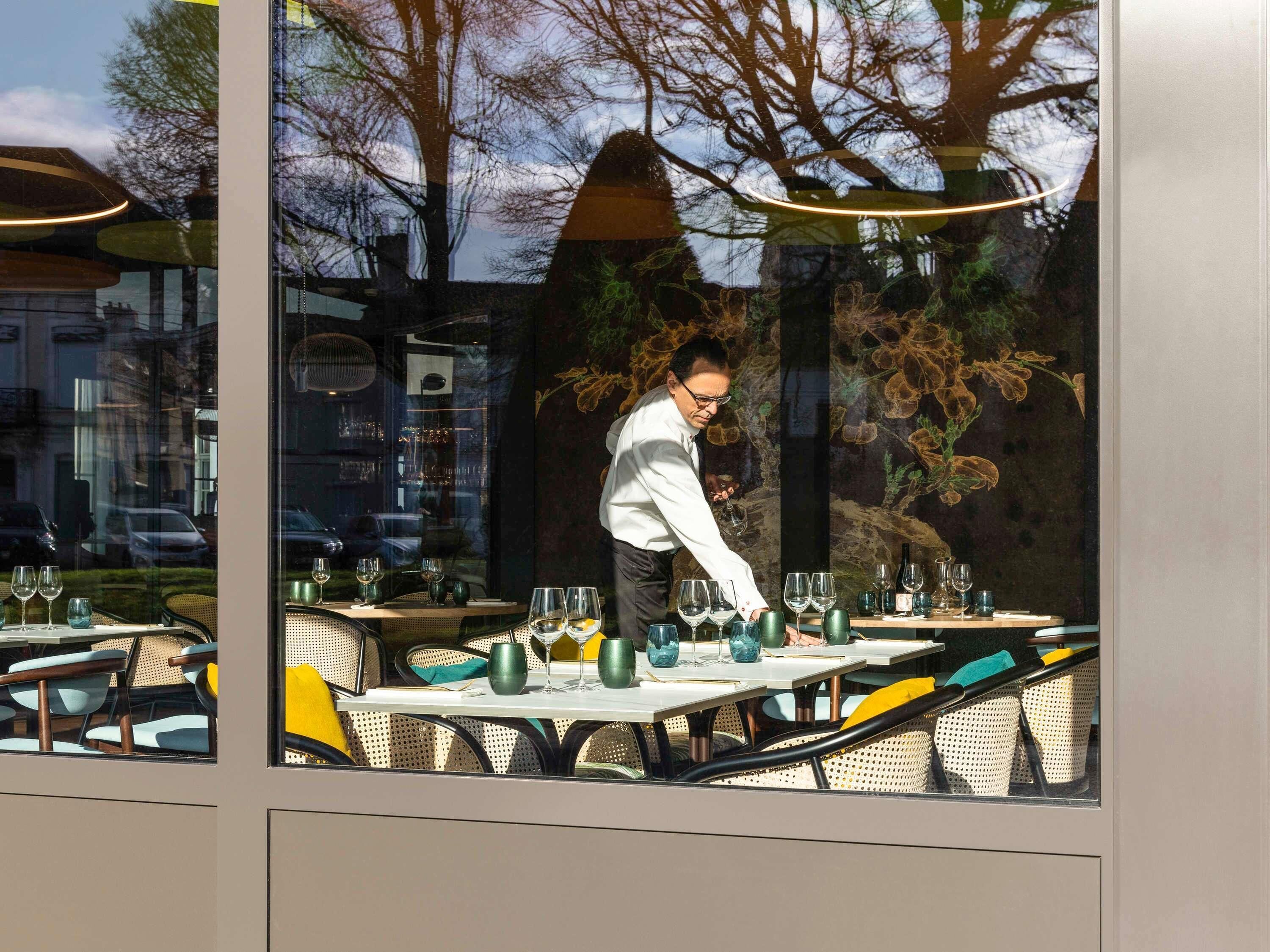 Un restaurant avec une terrasse, un serveur s’occupant d’une table, et une vue sur des arbres et un bâtiment en arrière-plan.