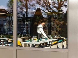 Un restaurante con terraza, un camarero atendiendo una mesa, y árboles y un edificio al fondo.