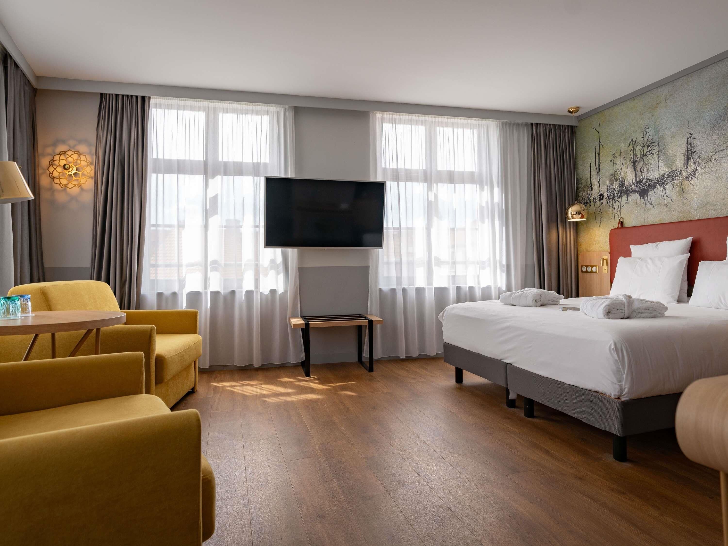 A hotel room with a bed, a TV, a desk with a chair, and a painting on the wall.