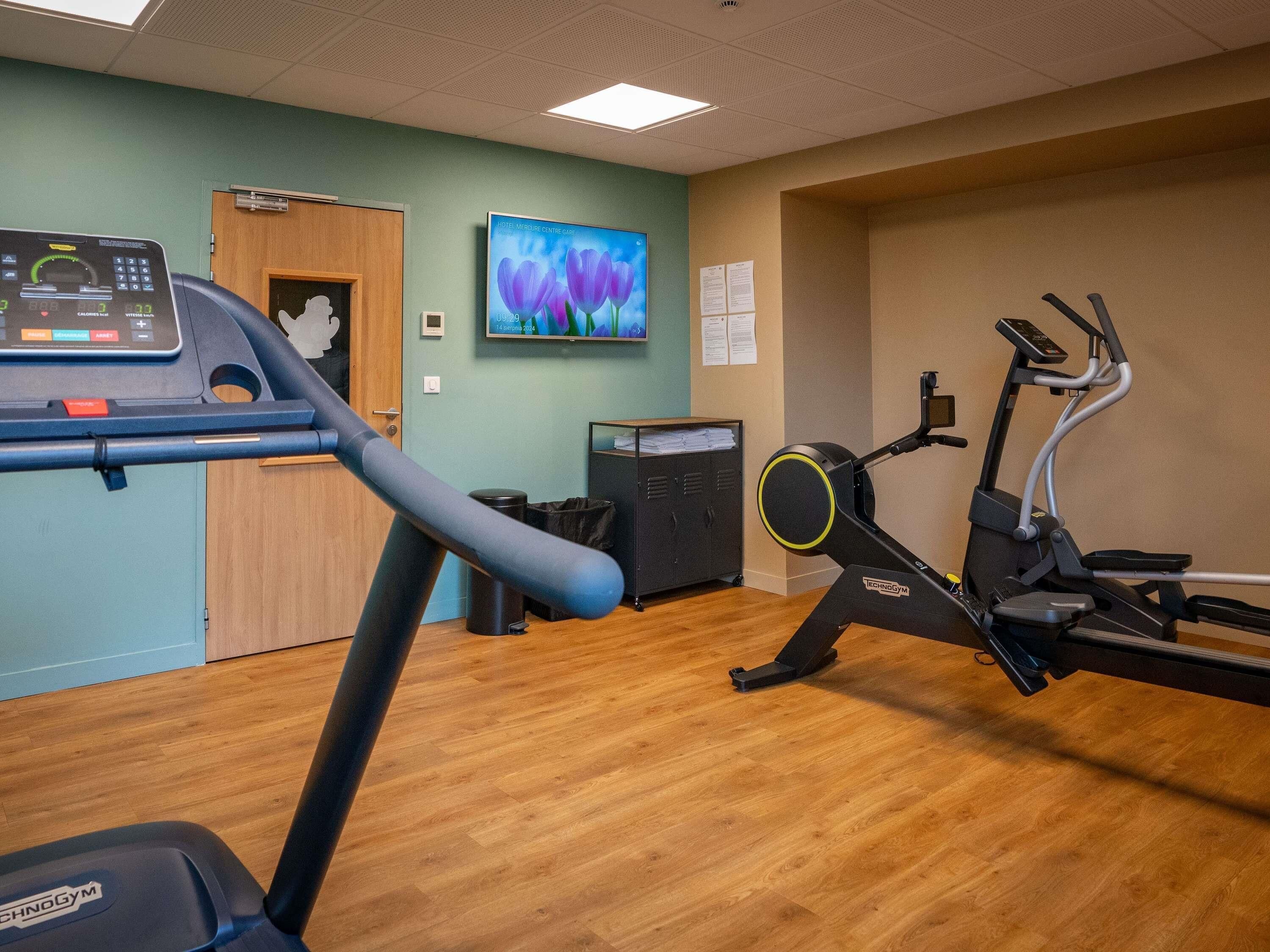 A fitness center with cardio equipment, a wooden floor, and a wall-mounted television.