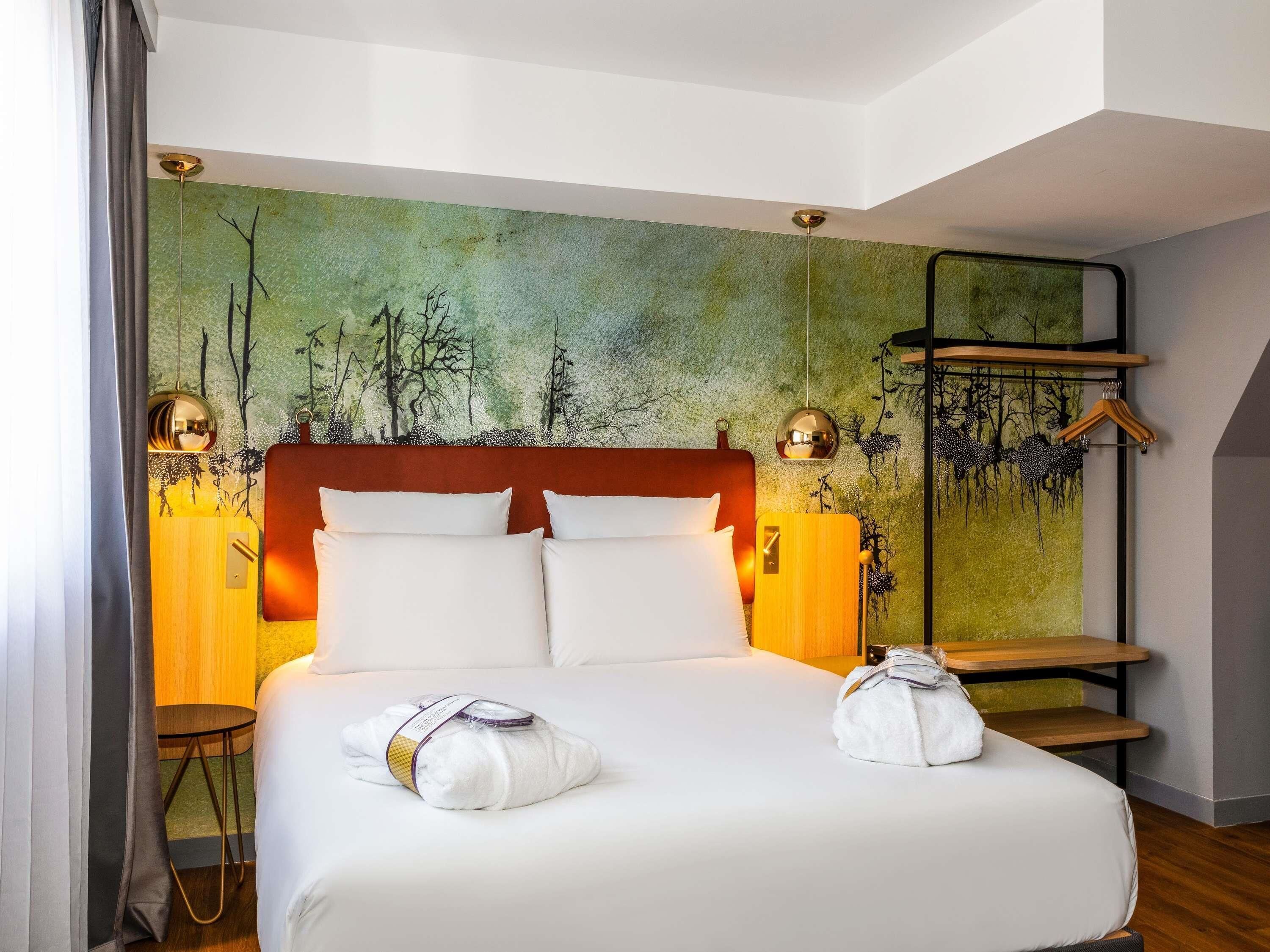 Habitació d'hotel amb un llit, un capçal, un mural a la paret, una taula de nit, una làmpada i un penjador.