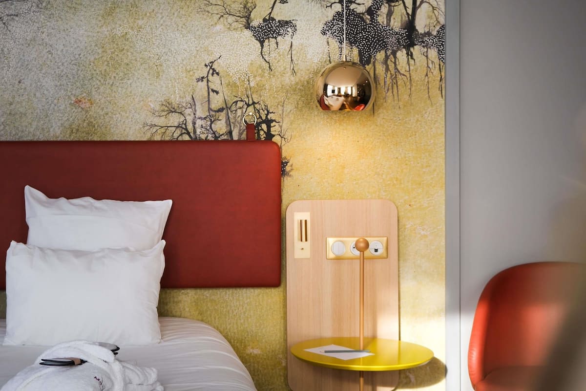 A hotel room with a bed, a wooden nightstand, a yellow table, and a red chair.