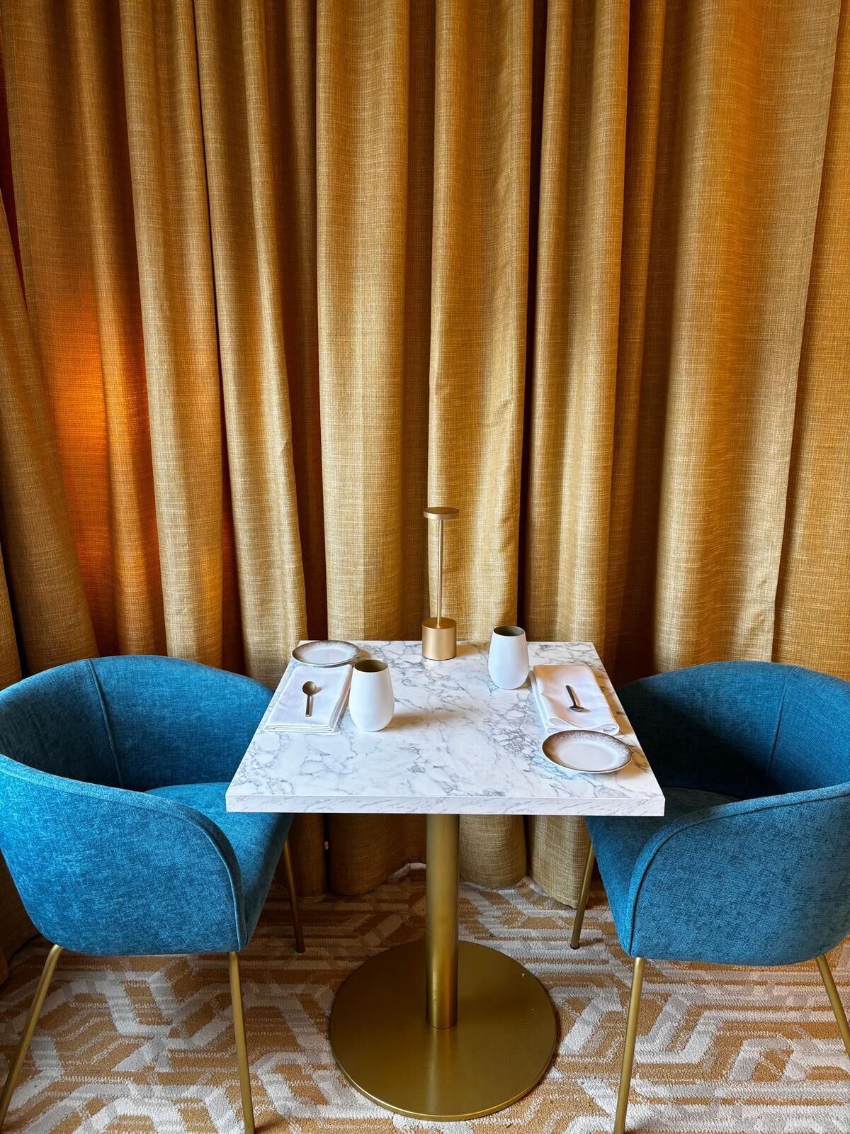 Una mesa de comedor con cubierta de mármol, dos sillas azules y una base dorada, frente a unos cortinajes dorados.