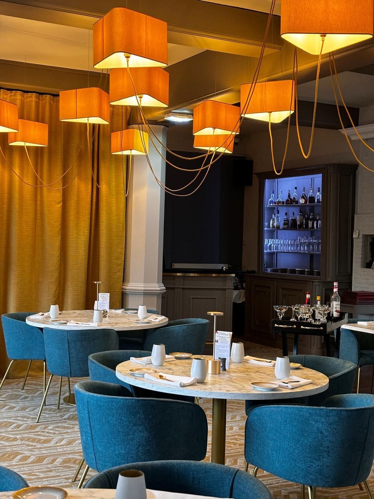 A hotel dining area with round tables, blue upholstered chairs, and pendant lighting.