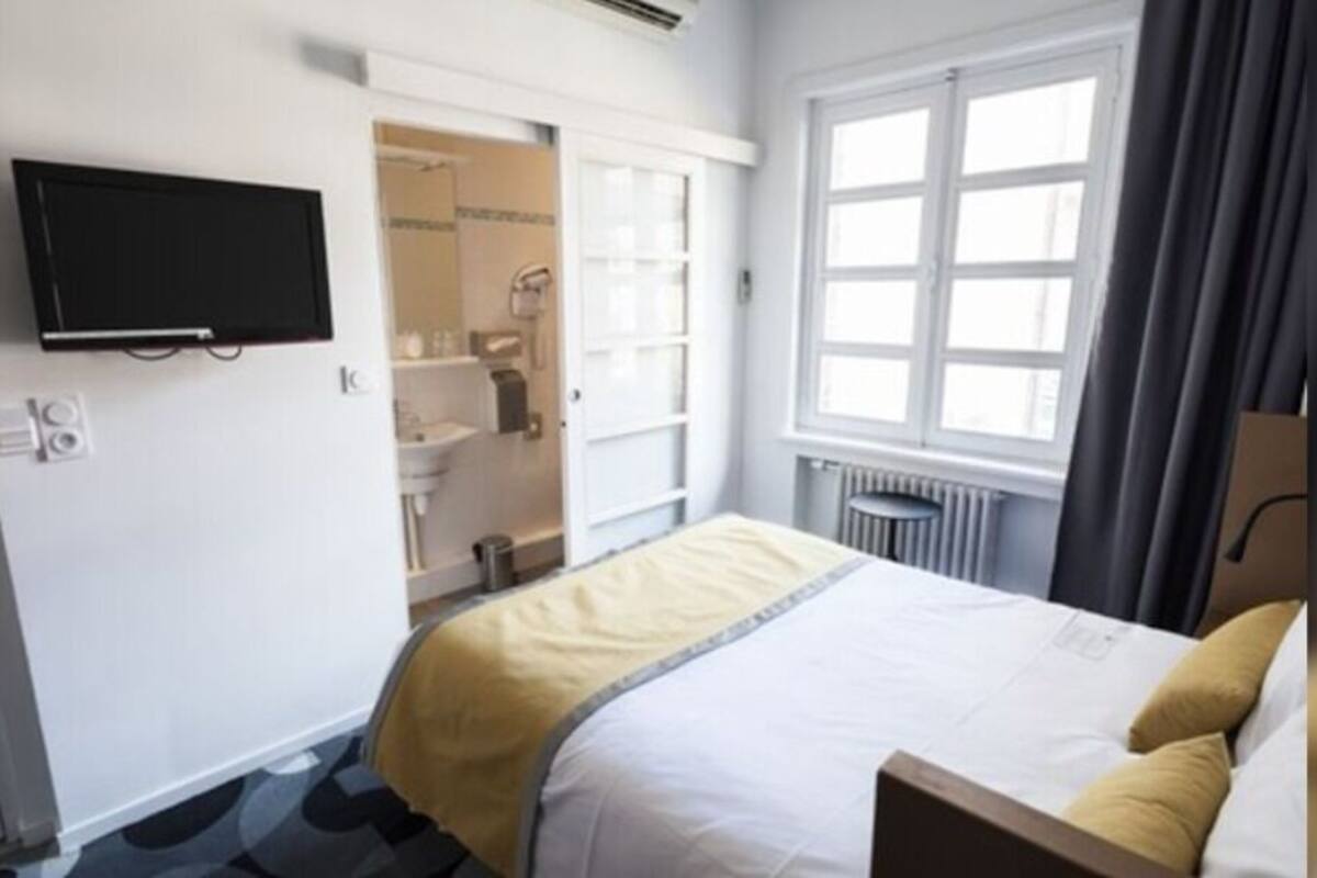 Ein Hotelzimmer mit Bett, Fernseher, einem Fenster mit Vorhängen, einer Heizung und einem sichtbaren Badezimmer durch eine offene Tür.