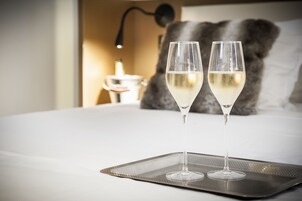 Dos copas de vino blanco sobre una bandeja en una cama, con una lámpara y una almohada grande al fondo.