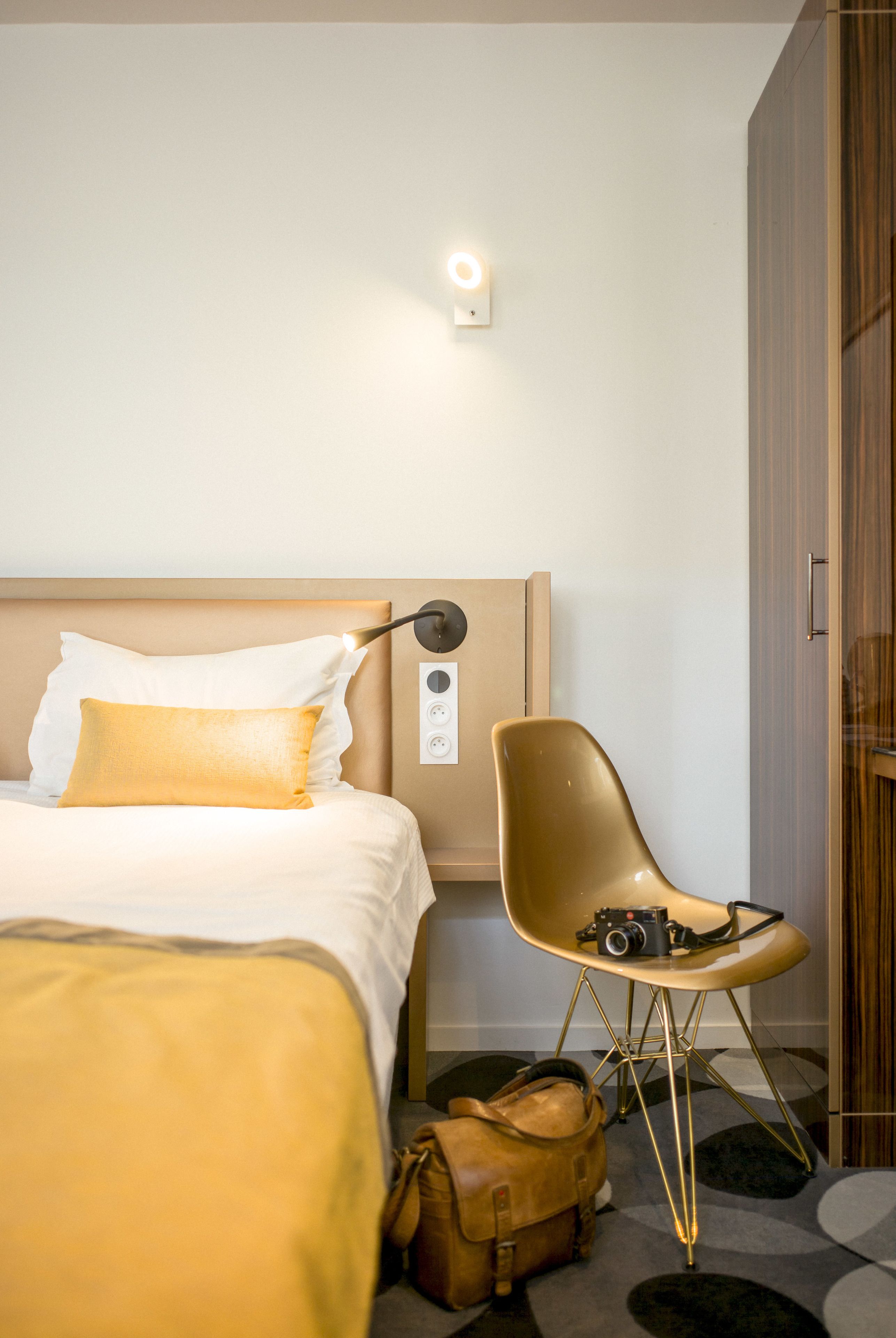 A hotel room with a bed, a chair, a camera, and a suitcase.