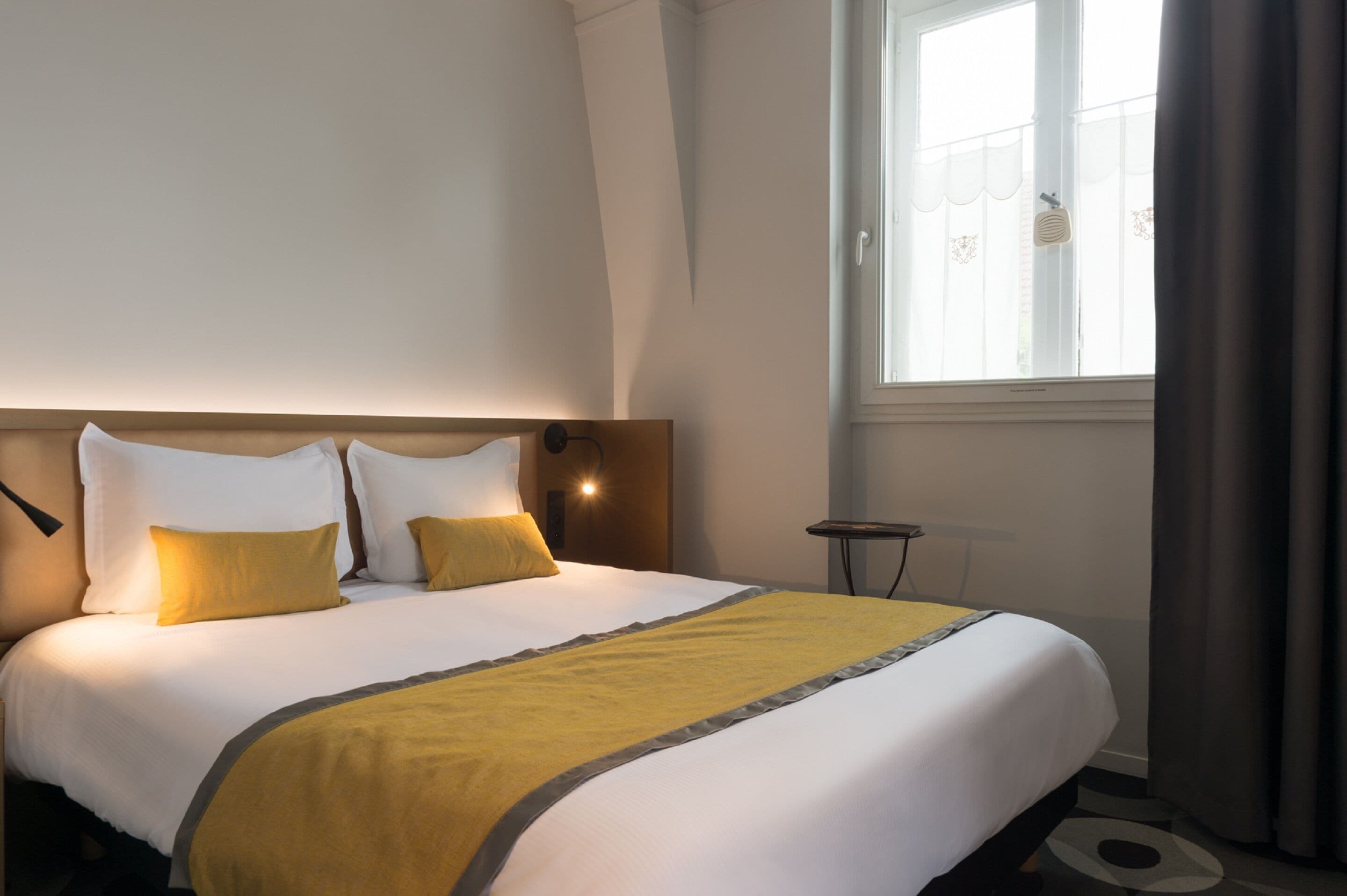 A neatly made bed with white and yellow bedding, a bedside table, and a window with curtains.