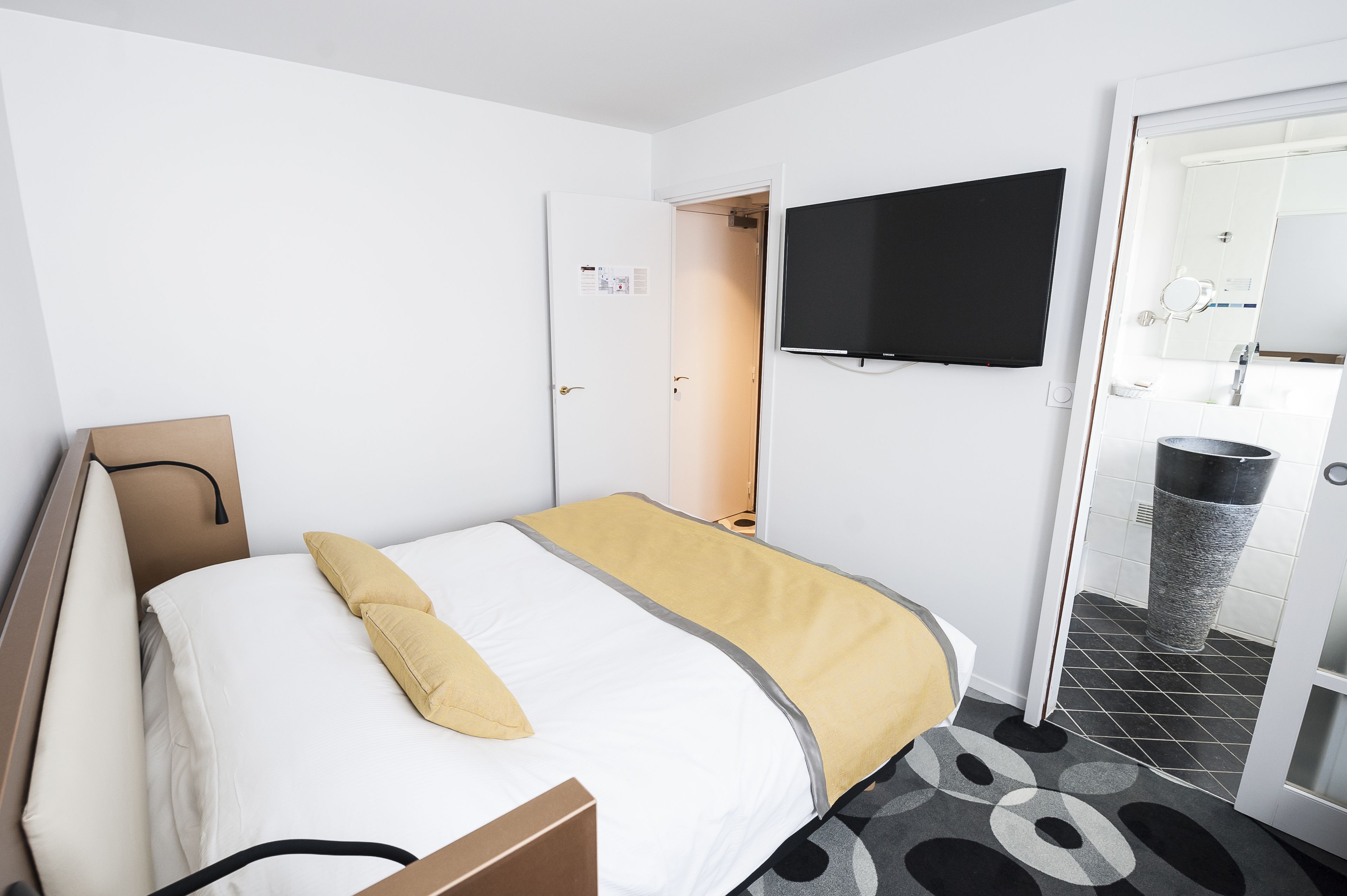 A hotel room with a bed, a television mounted on the wall, a bathroom with a mirror and sink, and a trash can.