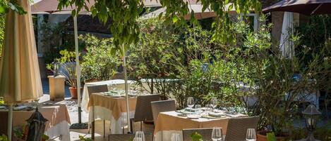 Un área de comedor al aire libre con mesas dispuestas para dos, rodeada de follaje y plantas en macetas.