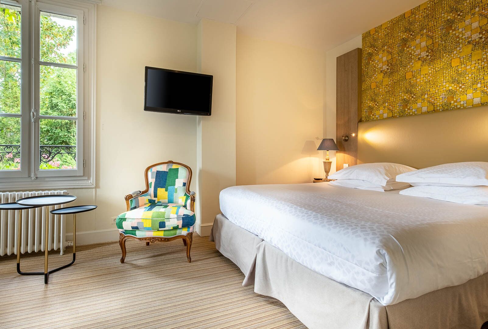 A hotel room with a bed, a chair, a TV, a lamp, and a window with a view of greenery.