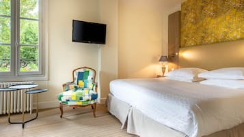A hotel room with a bed, a chair, a TV, a lamp, and a window with a view of greenery.