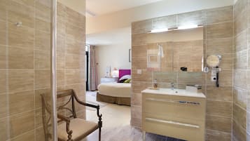 A modern bathroom with a wooden chair, a bed in the background, and a vanity with a mirror.