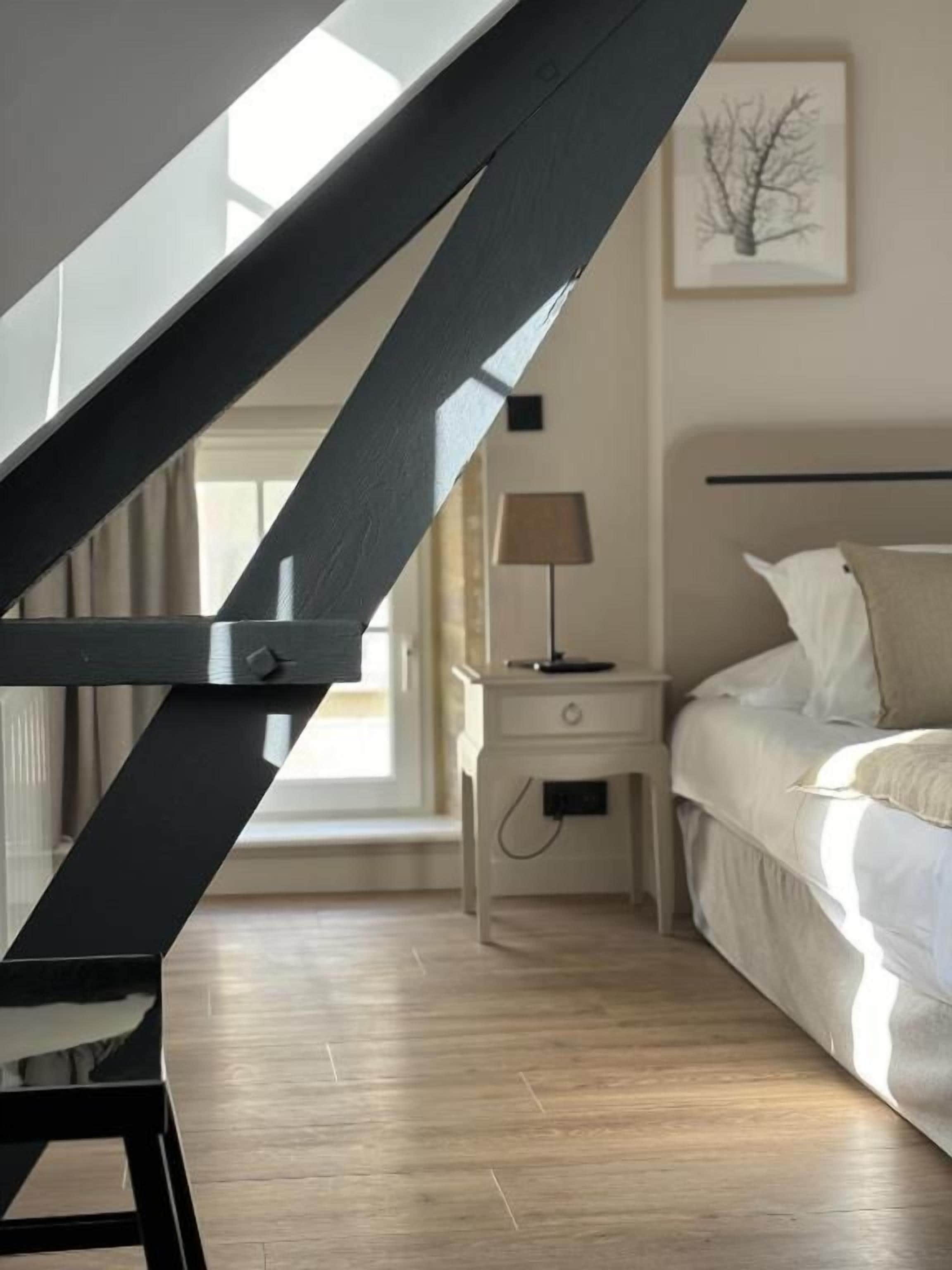 A bedroom with a bed, bedside table, lamp, and a view of a staircase.