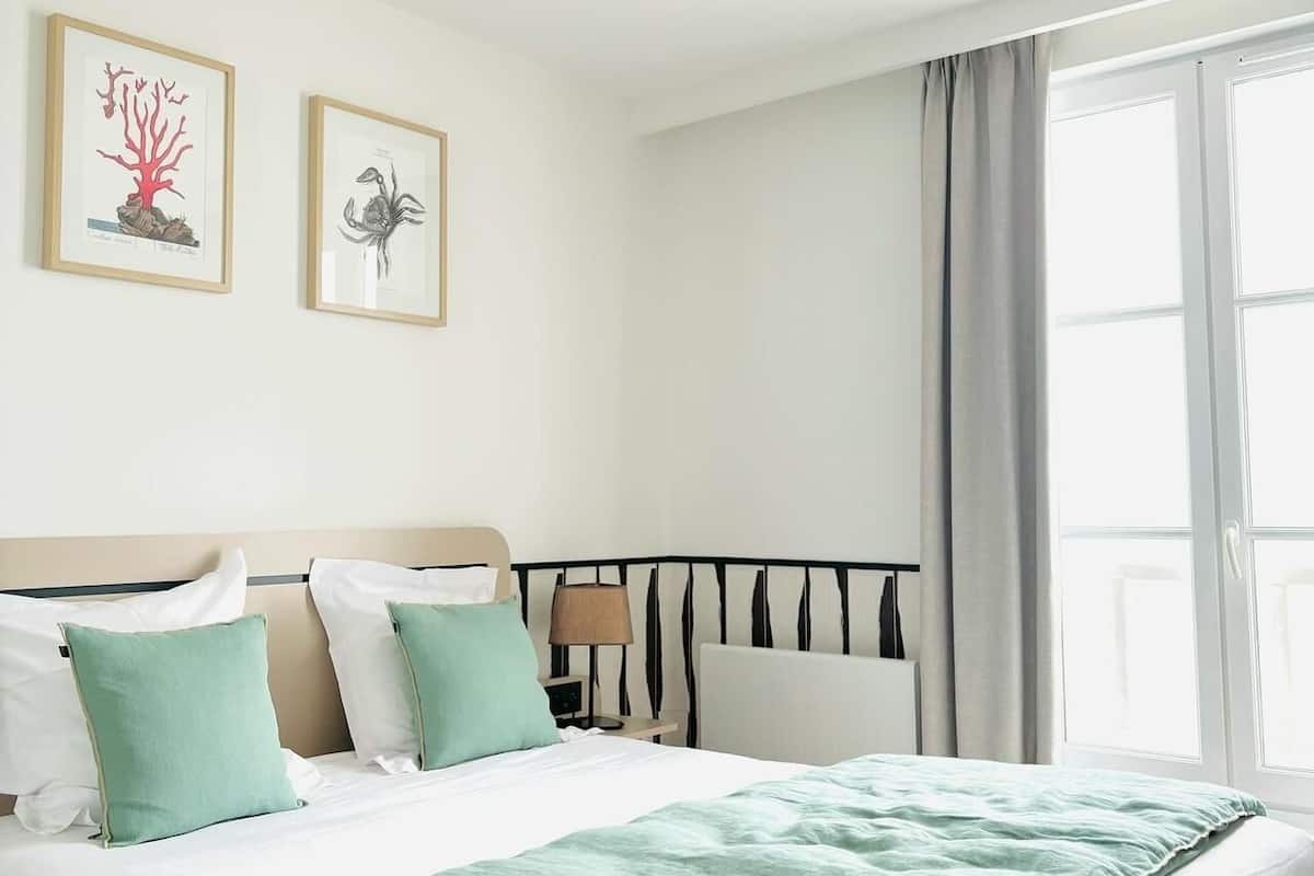 Une chambre d’hôtel moderne avec un grand lit, une tête de lit à lattes verticales, une fenêtre avec des rideaux et deux œuvres d’art encadrées accrochées au mur.