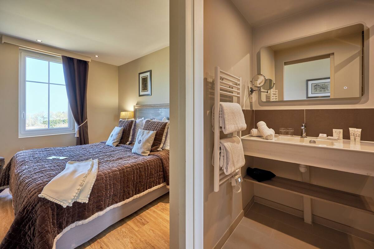 A hotel room with a bed, a window with curtains, a bedside table, and a bathroom with a sink, mirror, and towel rack.