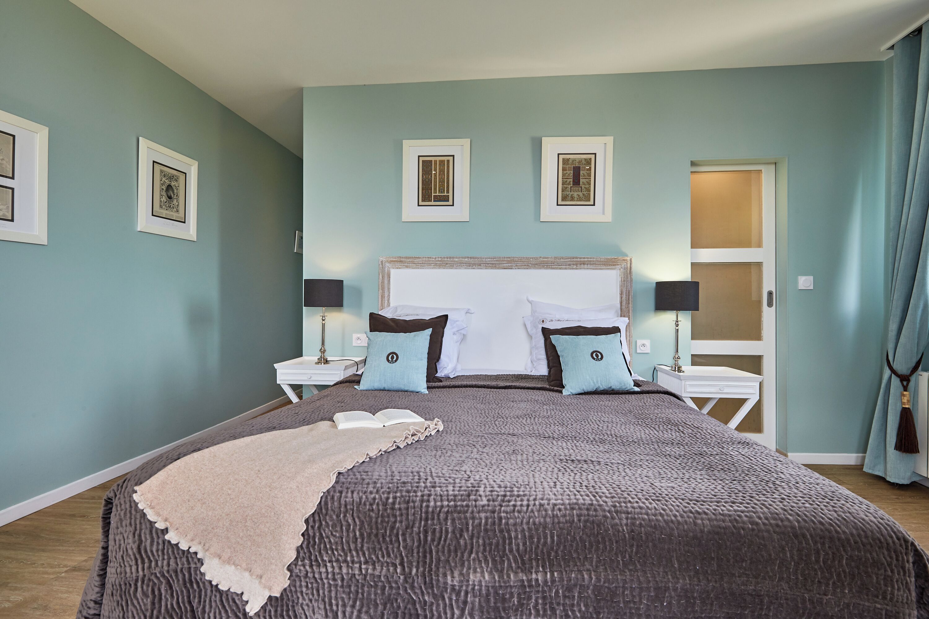 Une chambre à coucher avec un grand lit, deux tables de chevet avec des lampes, une fenêtre avec des rideaux et des œuvres d’art encadrées accrochées au mur.