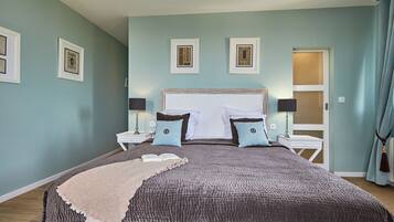 Une chambre à coucher avec un grand lit, deux tables de chevet avec des lampes, une fenêtre avec des rideaux et des œuvres d’art encadrées accrochées au mur.