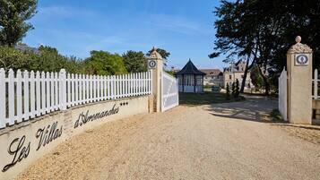 A gated entrance with a white picket fence, a sign reading "Les Villas d'Aromasches", and a building with a dark roof in the background.