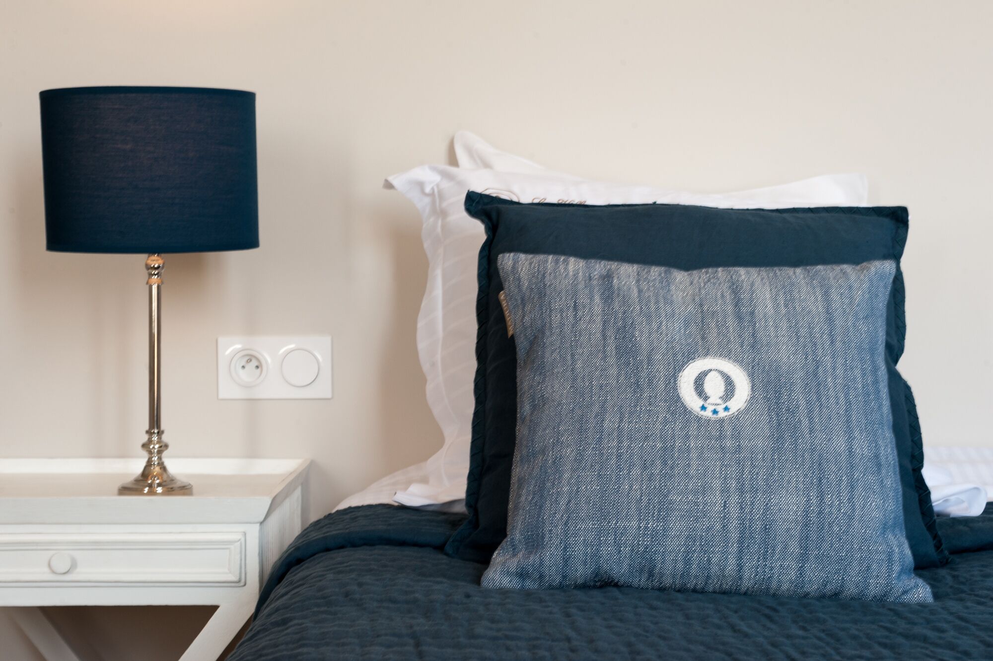 Een bed met een donkerblauwe sprei, een wit kussen en een donkerblauw kussen met een logo. Er staat een witte nachtkastje met een lamp en een stopcontact.