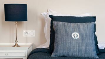 Een bed met een donkerblauwe sprei, een wit kussen en een donkerblauw kussen met een logo. Er staat een witte nachtkastje met een lamp en een stopcontact.