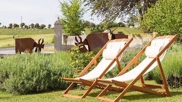 Dos sillones de madera en un área con césped, una pared de piedra y esculturas metálicas al fondo.