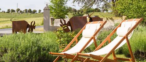 Dos sillones de madera en un área con césped, una pared de piedra y esculturas metálicas al fondo.