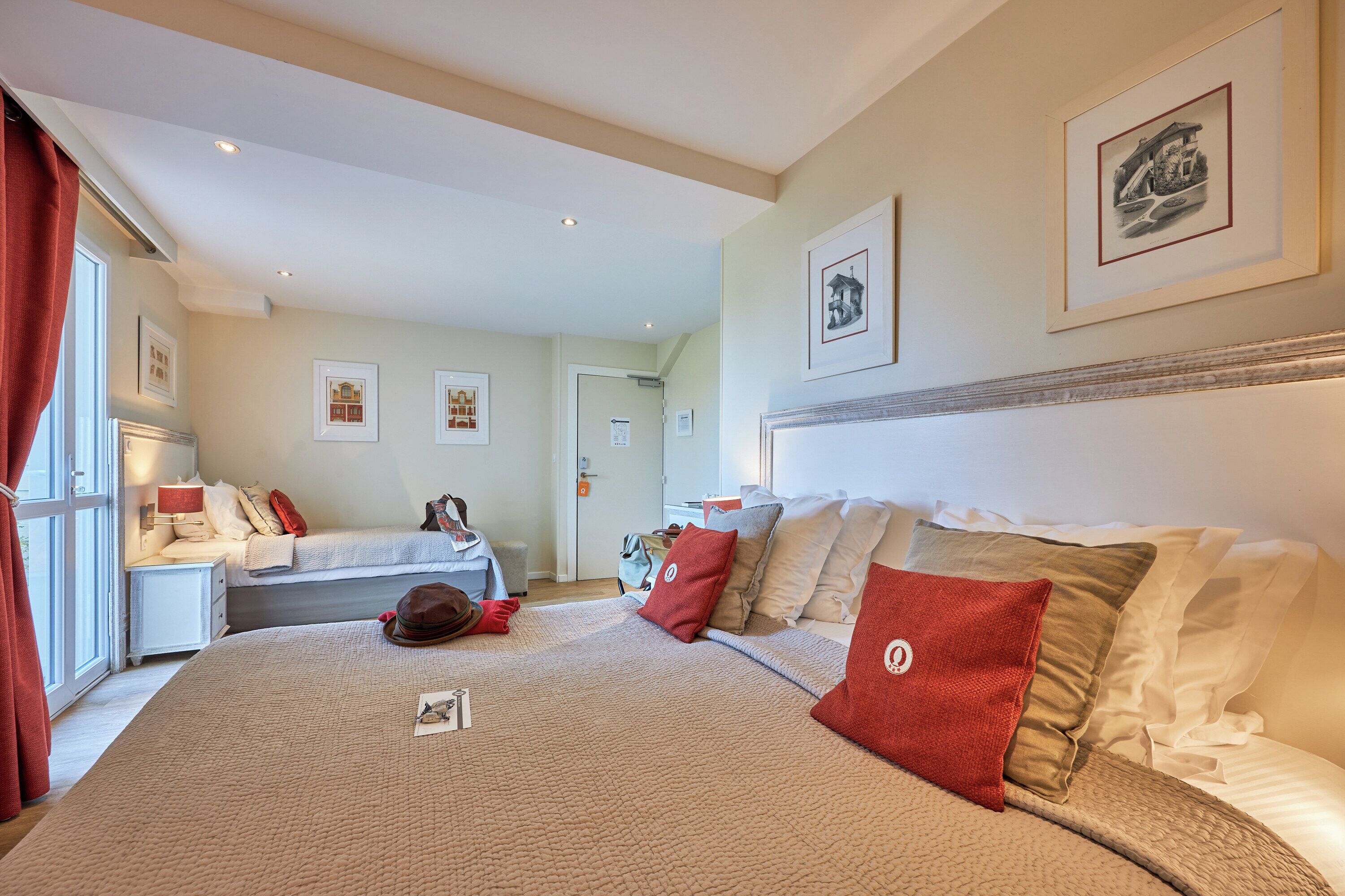 Een hotelkamer met een groot bed, een bureau en een stoel. Er hangen afbeeldingen in lijsten aan de muur en een raam met gordijnen.