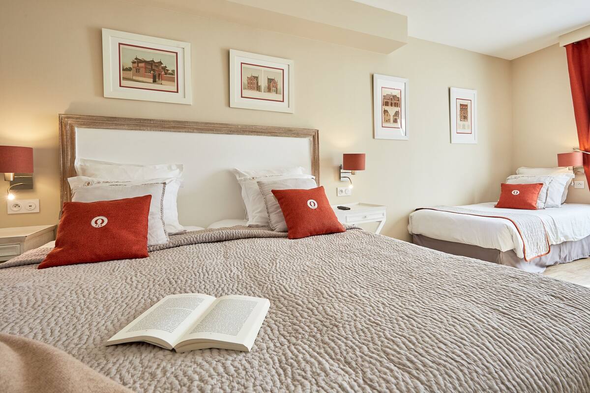 A hotel room with a large bed, two bedside tables, and a nightstand. There are framed pictures on the wall and a red pillow on the bed.