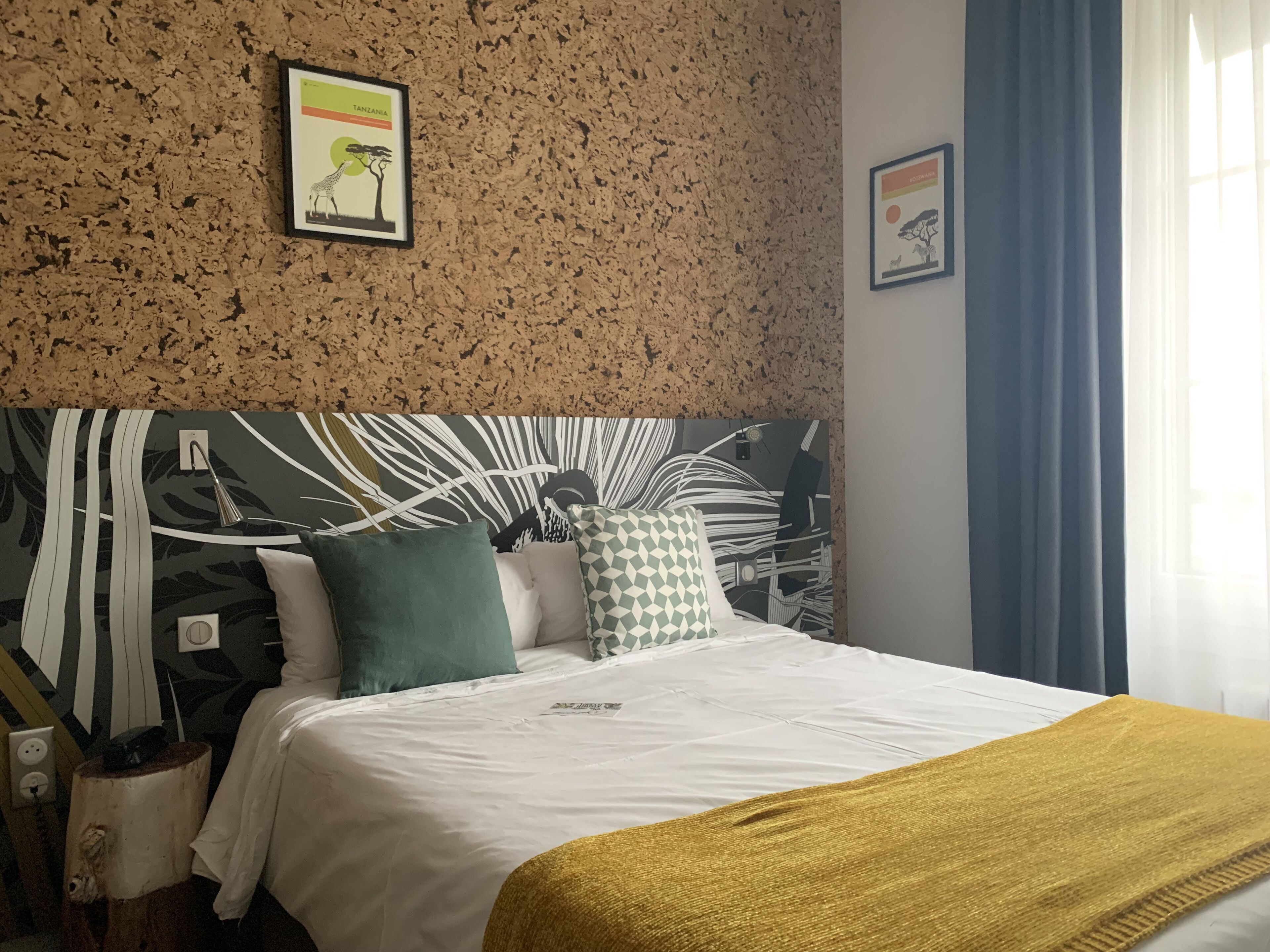 A hotel room with a bed, a headboard with a leaf pattern, a framed picture on the wall, and a window with curtains.