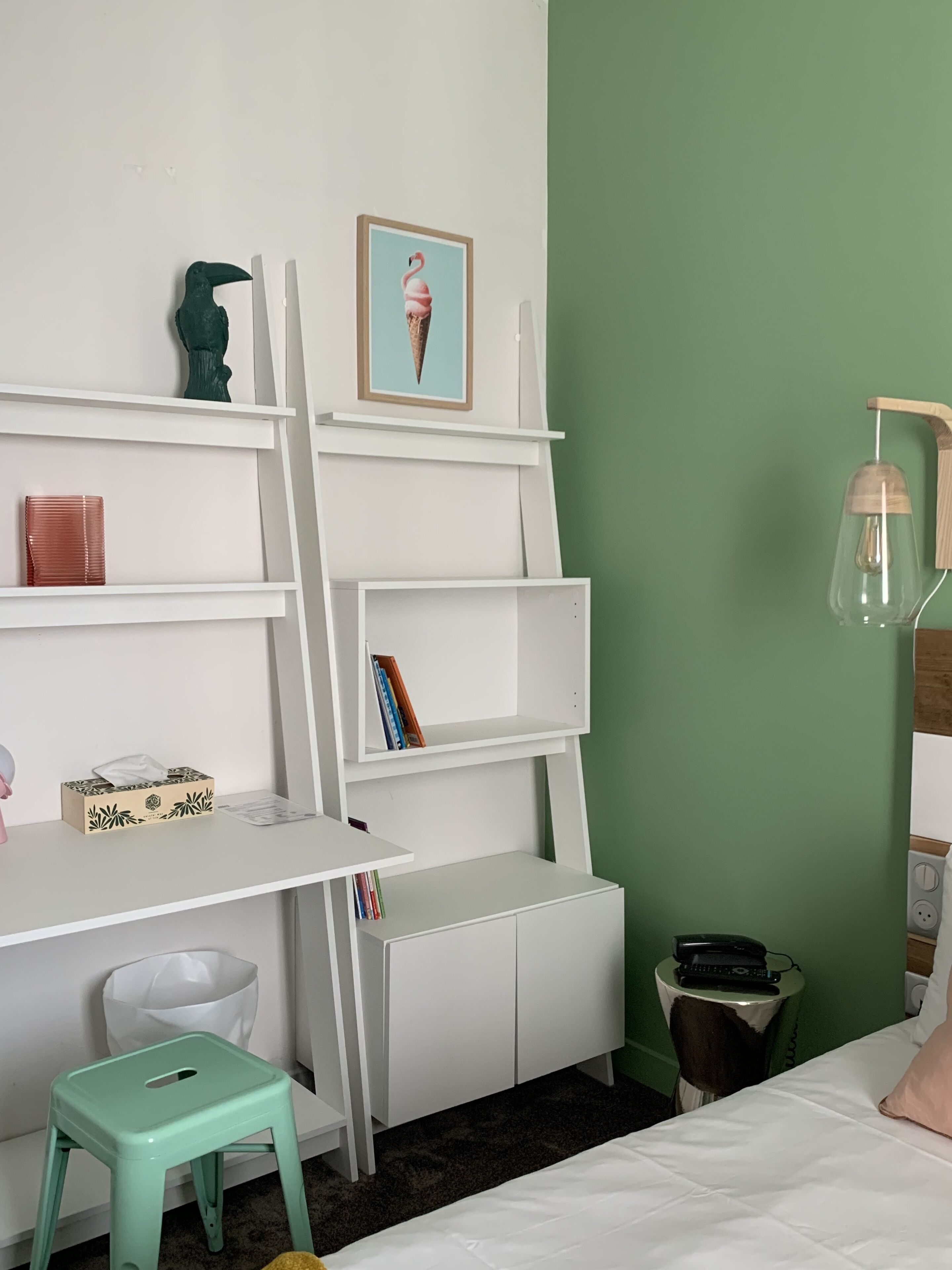 A room with a white desk, shelves, a small stool, a bed, and a green wall.