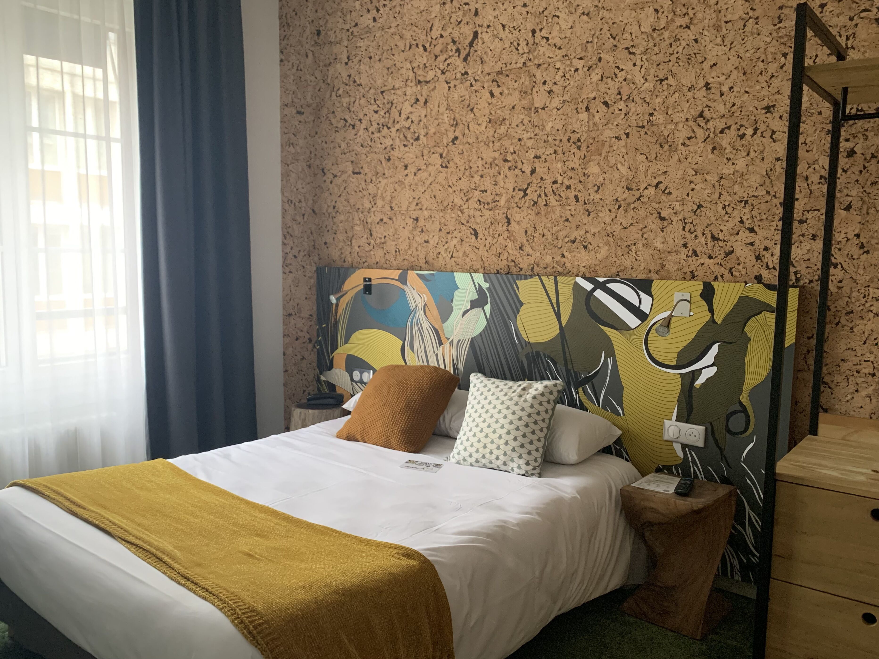 A neatly made bed with a white and mustard blanket, a patterned pillow, and a headboard with a colorful abstract design.