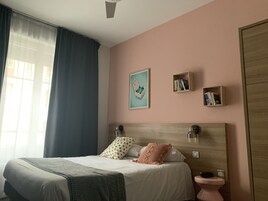 A bedroom with a bed, a bedside table, a nightstand, a window with curtains, a wall-mounted shelf with books, and a framed picture.