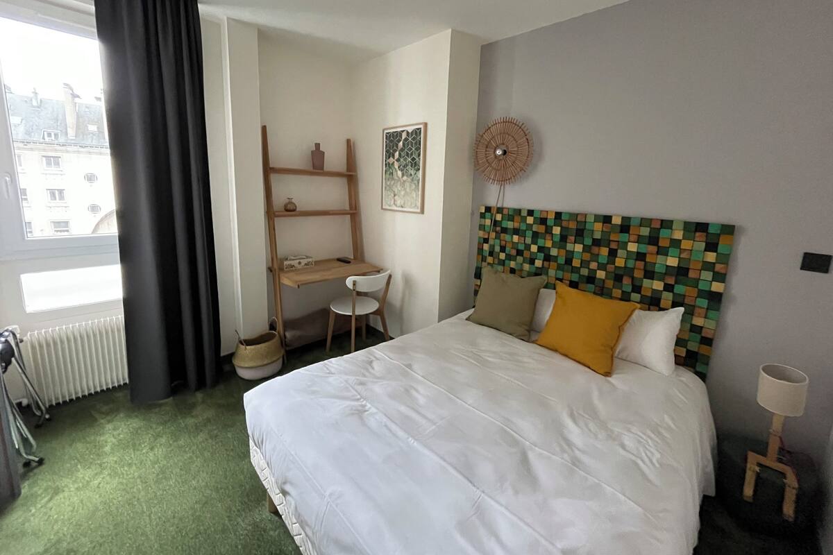 Ett hotellrum med en säng, ett skrivbord, en stol, ett element, ett fönster med gardiner och en väggmonterad fläkt.