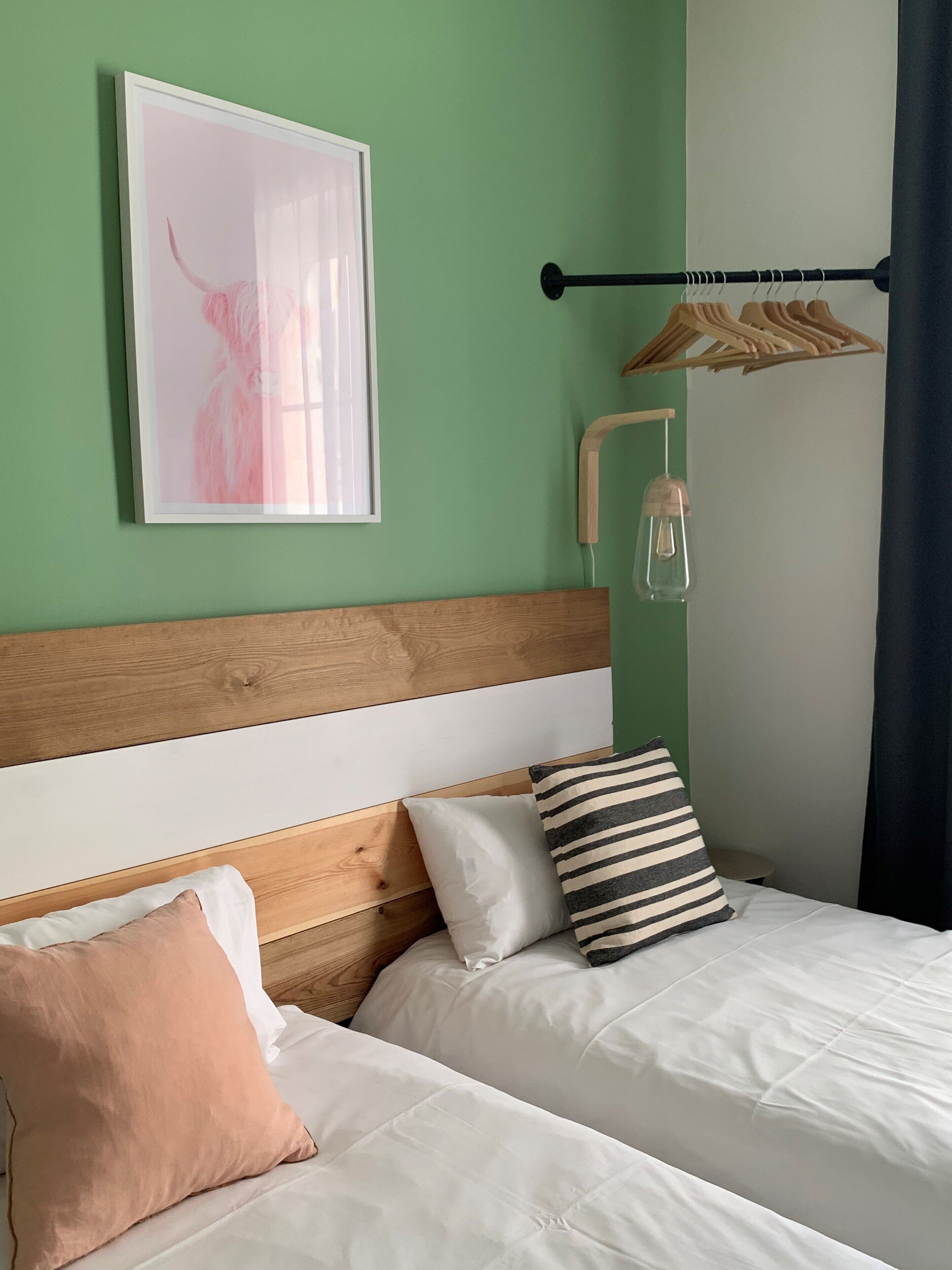Une chambre avec deux lits, une tête de lit en bois, un luminaire fixé au mur, un tableau encadré et un portant à vêtements.