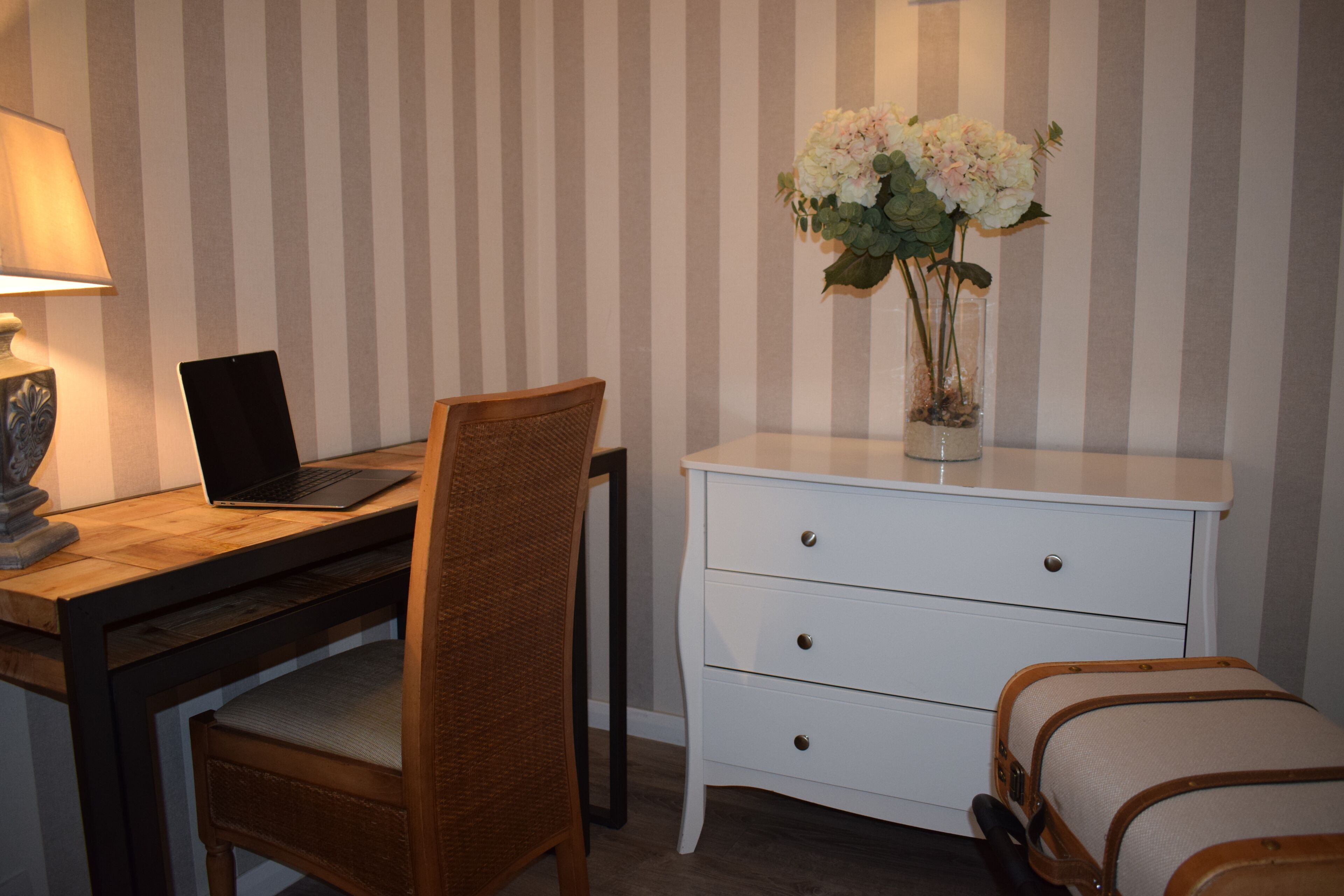 Et værelse med et træskrivebord, en bærbar computer, en fletstol, en hvid kommode med skuffer og en vase med blomster.