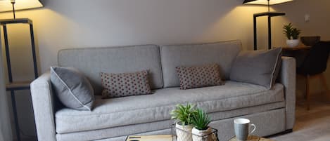 Una sala de estar moderna con un sofá gris, cojines estampados, dos lámparas de pie, una mesa de centro con una taza y una planta pequeña.
