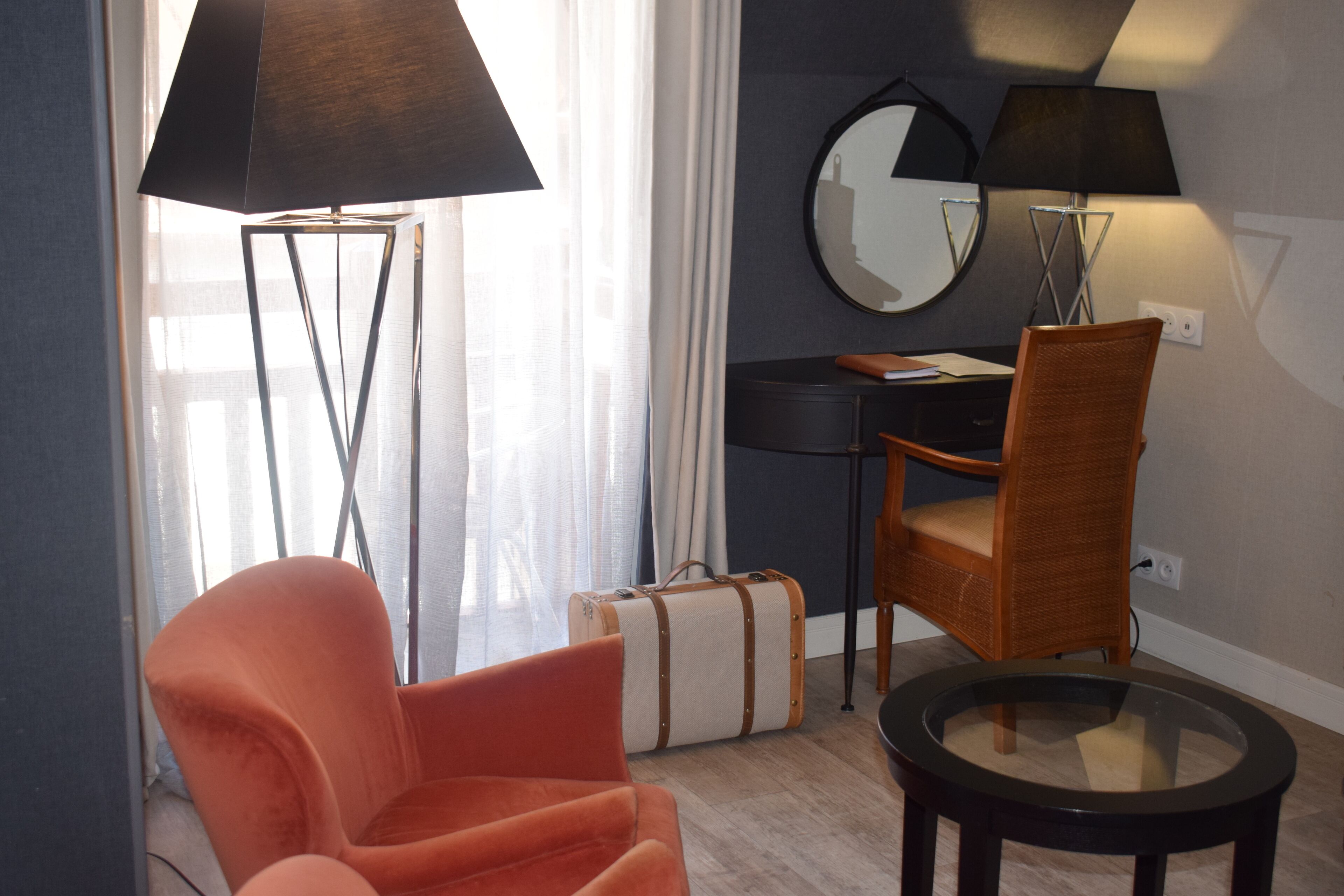 Una habitación con una silla moderna, una mesita, una lámpara de pie, una maleta y un escritorio con un espejo.