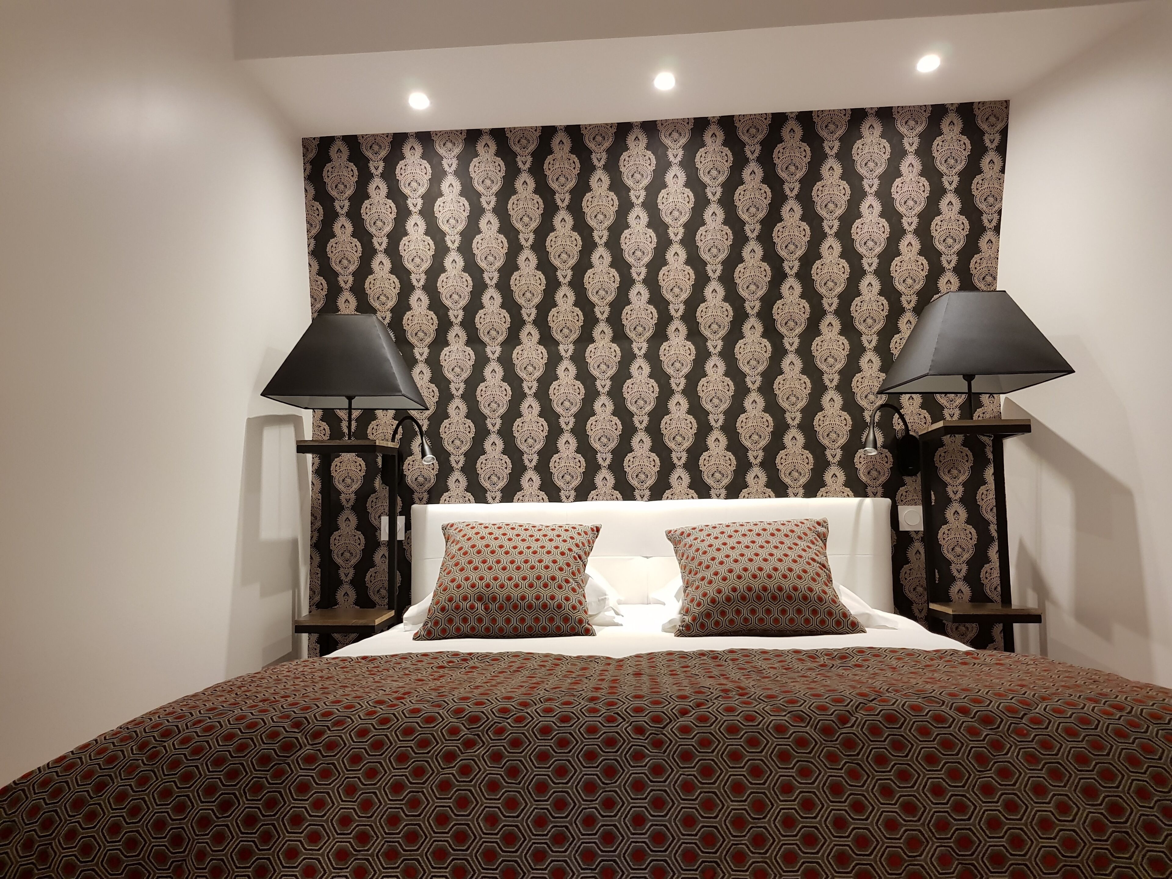 A neatly made bed with patterned bedding, two bedside lamps, and a patterned wallpaper.