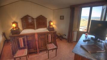 A bedroom with a wooden bed, two bedside lamps, a desk with a computer, and a view of the landscape.