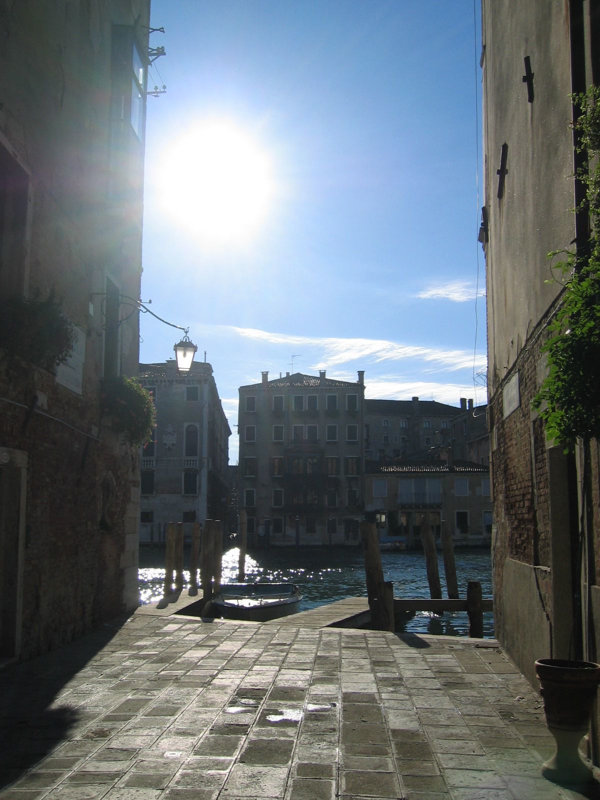 A narrow Venetian canal with buildings on either side, a clear sky, and a bright sun shining through.