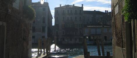 A narrow Venetian canal with buildings on either side, a clear sky, and a bright sun shining through.