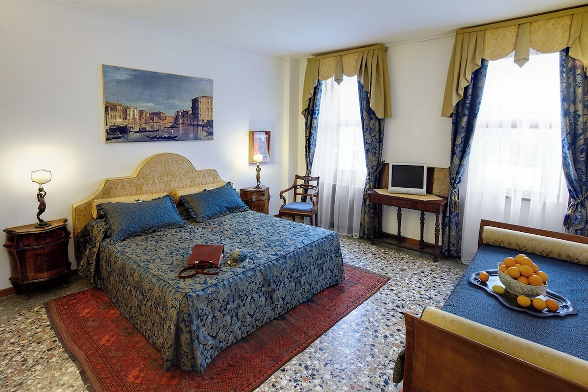 A hotel room with a bed, a desk with a computer, a chair, a TV, and a tray of oranges.