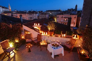 Une terrasse extérieure avec une table dressée pour le dîner, entourée de chaises et de bougies, d’où l’on peut admirer un panorama sur la ville au crépuscule.