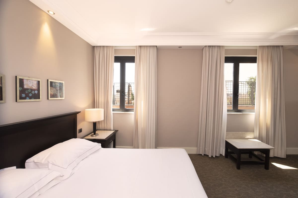 Een hotelkamer met een groot bed, een nachtkastje met een lamp, een klein tafeltje en twee ramen met gordijnen.