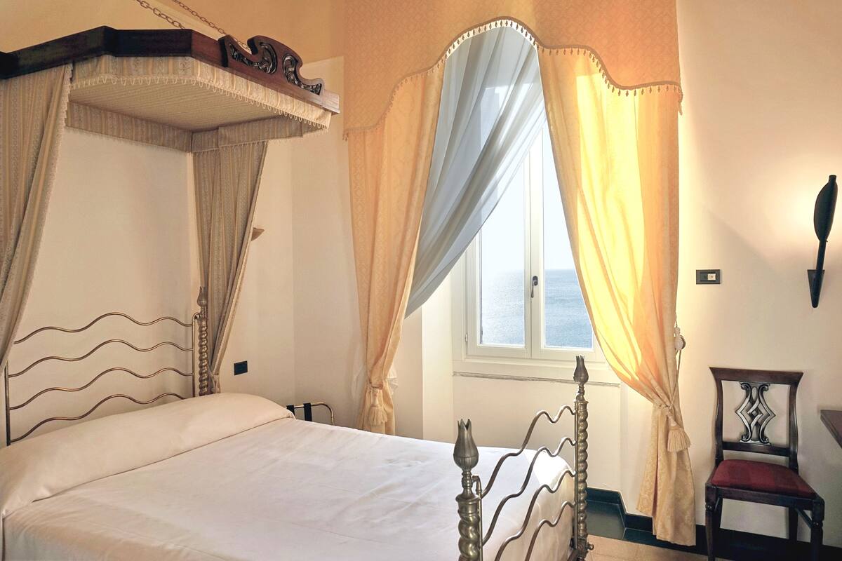 Una cama con dosel, una silla y un ventanal con cortinas.