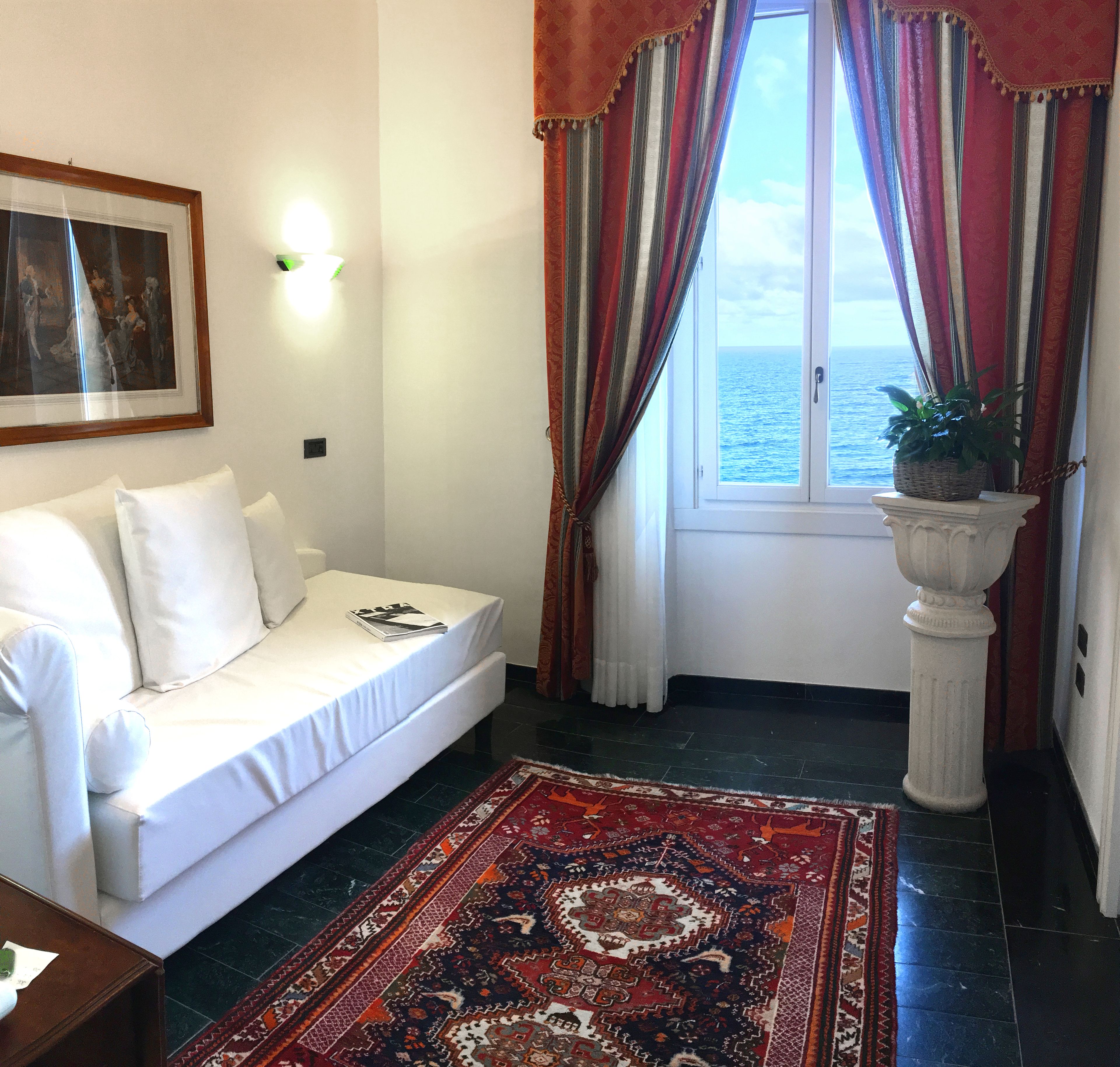 Una stanza con un divano bianco, un quadro appeso al muro, una pianta in vaso su un piedistallo e vista sul mare attraverso una finestra con tende a righe.