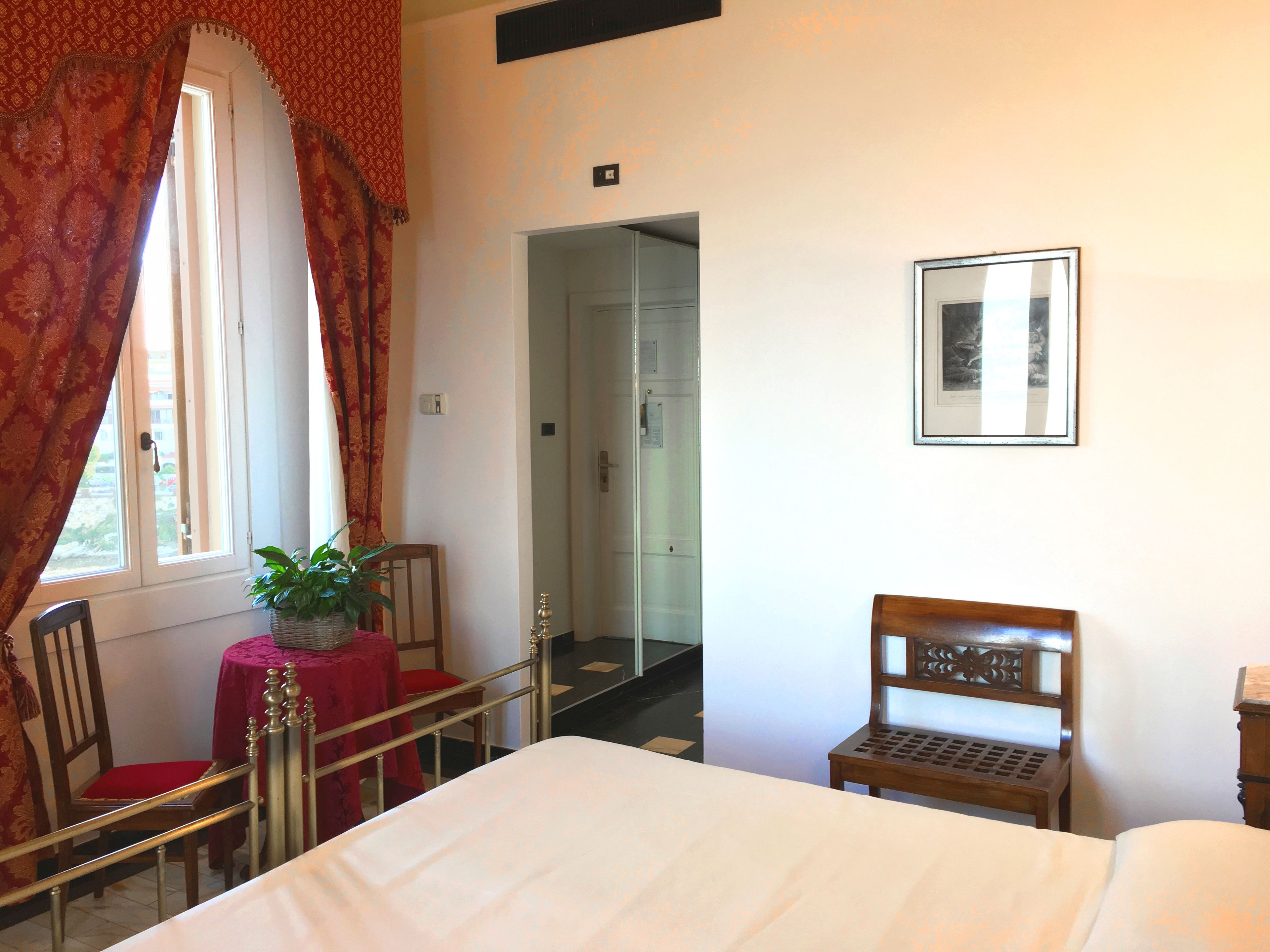 Habitación de hotel con una cama, una silla de madera, una mesita con una planta, una silla roja, una ventana con cortinas y una puerta que da a otra habitación.